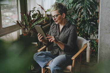 Mature woman using tablet PC sitting on chair in front of plants at home - YTF00543