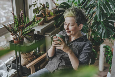 Nachdenkliche reife Frau beim Kaffee inmitten von Zimmerpflanzen zu Hause - YTF00547