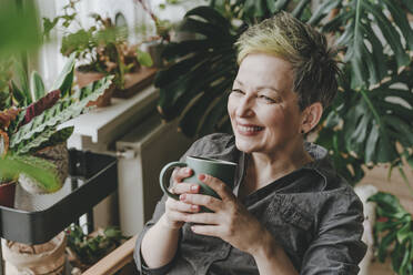 Lächelnde Frau sitzt mit Kaffeetasse inmitten von Topfpflanzen zu Hause - YTF00548