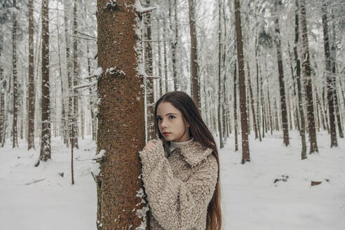 Nachdenkliches Teenager-Mädchen steht an einem Baum im verschneiten Wald - VSNF00519
