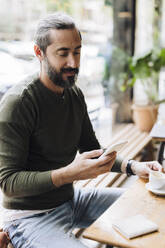 Mann mit Kaffeetasse und Smartphone am Schreibtisch in einem Cafe - JOSEF17161