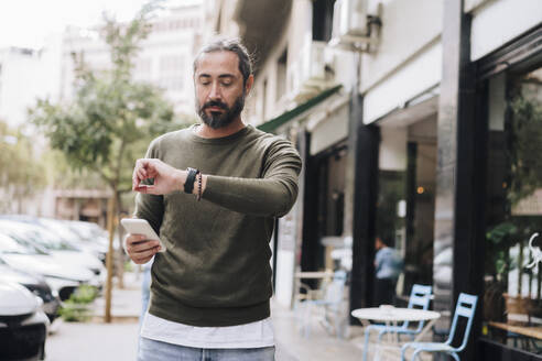 Mann hält Smartphone und prüft die Zeit auf der Armbanduhr - JOSEF17169