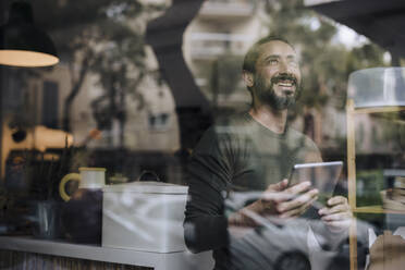 Glücklicher reifer Mann mit Tablet-PC im Café sitzend durch Glas gesehen - JOSEF17184