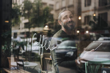 Lächelnder reifer Mann mit Tablet-PC im Café sitzend durch Glas gesehen - JOSEF17199