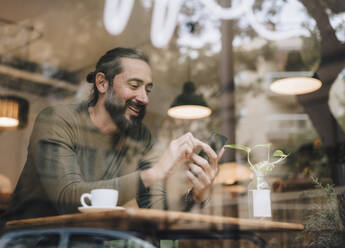 Smiling mature man using smart phone sitting at table in cafe - JOSEF17214