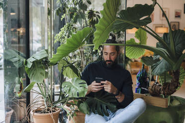 Mann mit Mobiltelefon inmitten von Pflanzen sitzend - JOSEF17237