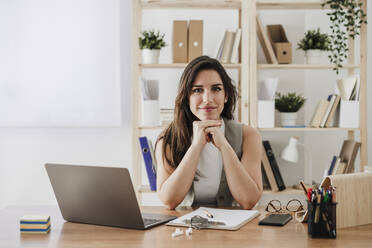 Smiling businesswoman sitting with hands on chin at desk in office - EBBF08039