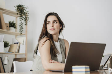 Thoughtful businesswoman sitting with laptop in office - EBBF08042