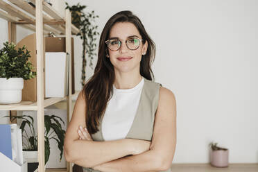 Confident smiling businesswoman standing with arms crossed by shelf in office - EBBF08051