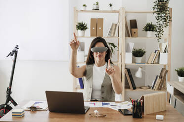Businesswoman wearing VR glasses sitting at desk in office - EBBF08061