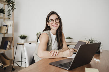 Happy businesswoman sitting in front of laptop at office desk - EBBF08076