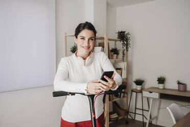 Smiling businesswoman standing with electric scooter holding smart phone in office - EBBF08086