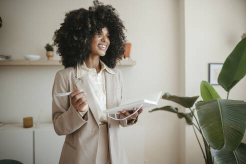 Happy businesswoman with Afro hairstyle standing with book in office - RCPF01669