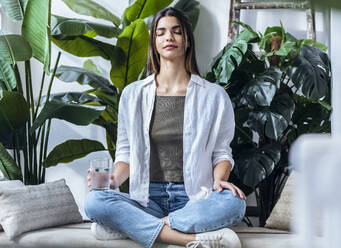 Young woman meditating on sofa in front of green plants - JSRF02377