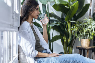 Junge Frau trinkt Wasser bei Pflanzen sitzend - JSRF02378
