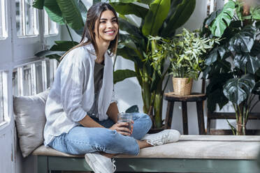 Smiling young woman with glass of water sitting on couch - JSRF02379