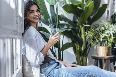 Happy young woman drinking glass of water at the window - JSRF02380