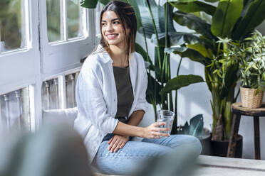 Happy young woman sitting with glass of water at the window - JSRF02382