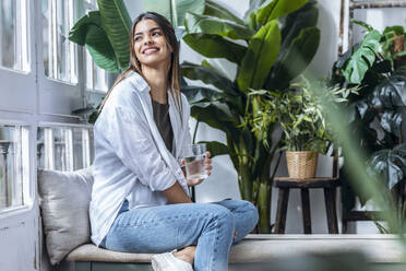 Happy woman sitting with glass of water at the window - JSRF02384