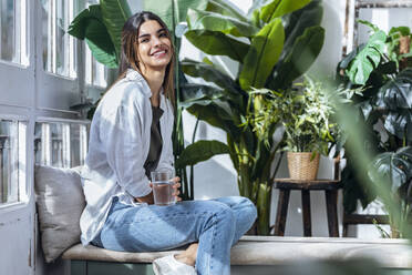 Happy woman sitting with glass of water at the window - JSRF02386