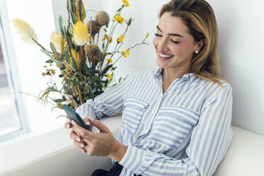 Smiling woman using mobile phone sitting on sofa - JSRF02398