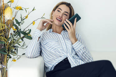 Happy young woman talking through speaker phone on sofa - JSRF02403