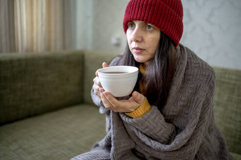 In eine Decke eingewickelte Frau sitzt mit einer Tasse Tee zu Hause - ANAF01018