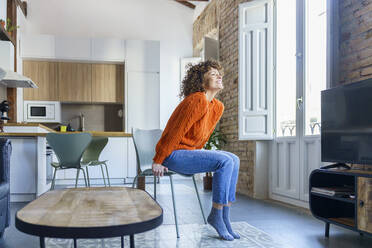 Happy curly haired woman sitting on chair at home - JSMF02672