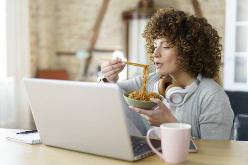 Freelancer with bowl of noodles working on laptop at home - JSMF02699