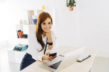 Lächelnde Freiberuflerin mit Mobiltelefon und Laptop am Schreibtisch sitzend - JJF00225
