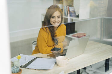 Happy female freelancer with smart phone and laptop sitting at desk - JJF00237