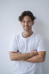 Happy young man with arms crossed standing in front of wall - KNSF09658