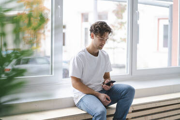 Junger Mann mit Smartphone vor einem Fenster sitzend - KNSF09663