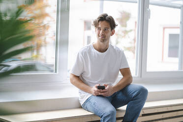Smiling young man sitting with smart phone in front of window - KNSF09664