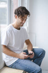 Happy young man using smart phone by window - KNSF09665
