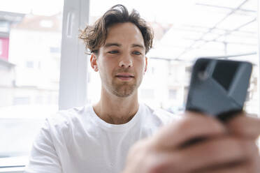 Junger Mann mit Mobiltelefon vor einem Fenster - KNSF09668