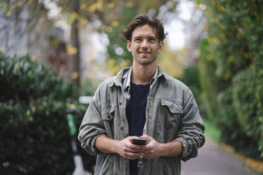 Thoughtful young man standing with smart phone - KNSF09681