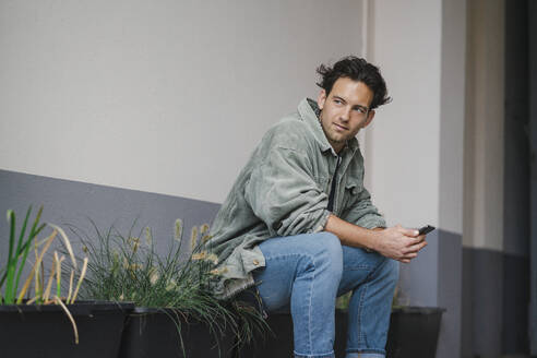 Junger Mann sitzt bei Topfpflanzen mit Smartphone - KNSF09682