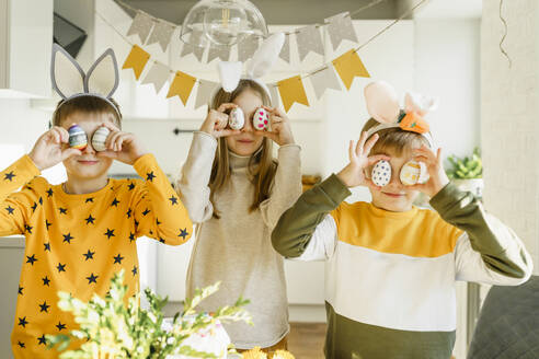 Smiling boys and girl holding Easter eggs over eyes at home - VBUF00265