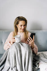 Lächelnde junge Frau mit Smartphone und Kaffeetasse zu Hause - JJF00244