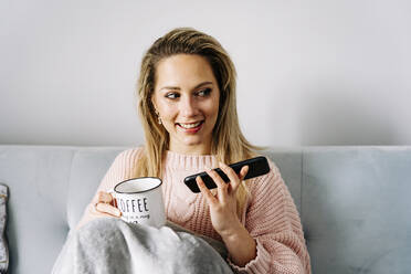 Glückliche Frau mit Smartphone und Kaffeetasse auf dem Sofa zu Hause - JJF00245