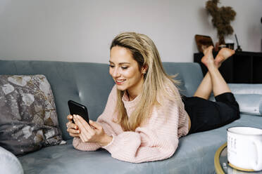 Smiling young woman using smart phone lying on sofa at home - JJF00250