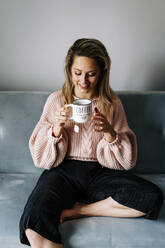 Happy woman enjoying coffee sitting on sofa at home - JJF00253