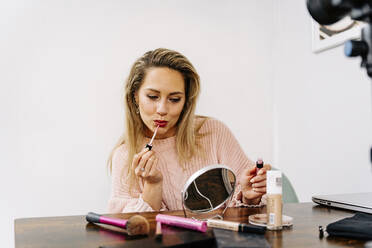 Young woman looking in mirror applying lipstick at table - JJF00258