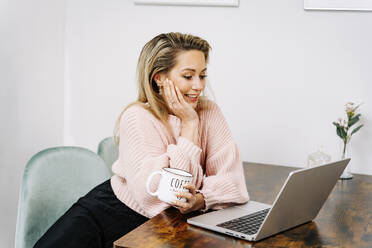 Happy woman with coffee cup using laptop at home - JJF00260