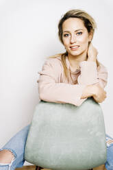 Young woman sitting on hair in front of white wall - JJF00266