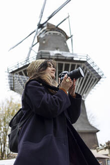 Young woman photographing through camera in front of windmill - AXHF00267