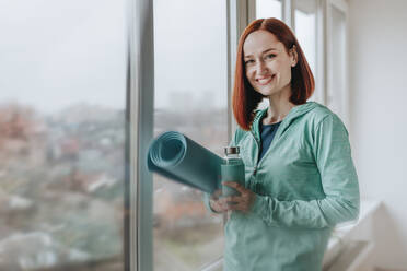 Lächelnde Frau mit Übungsmatte und Wasserflasche in der Nähe des Fensters stehend - YTF00549
