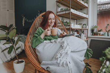 Lächelnde Frau mit einer Tasse Kaffee und einem Tablet-PC auf einem Stuhl zu Hause - YTF00563