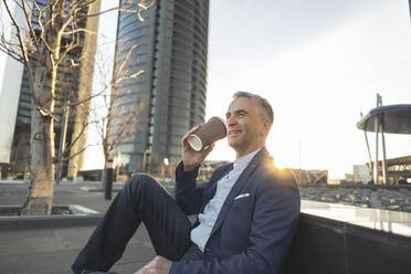 Smiling businessman with coffee cup sitting at office park - JCCMF09460